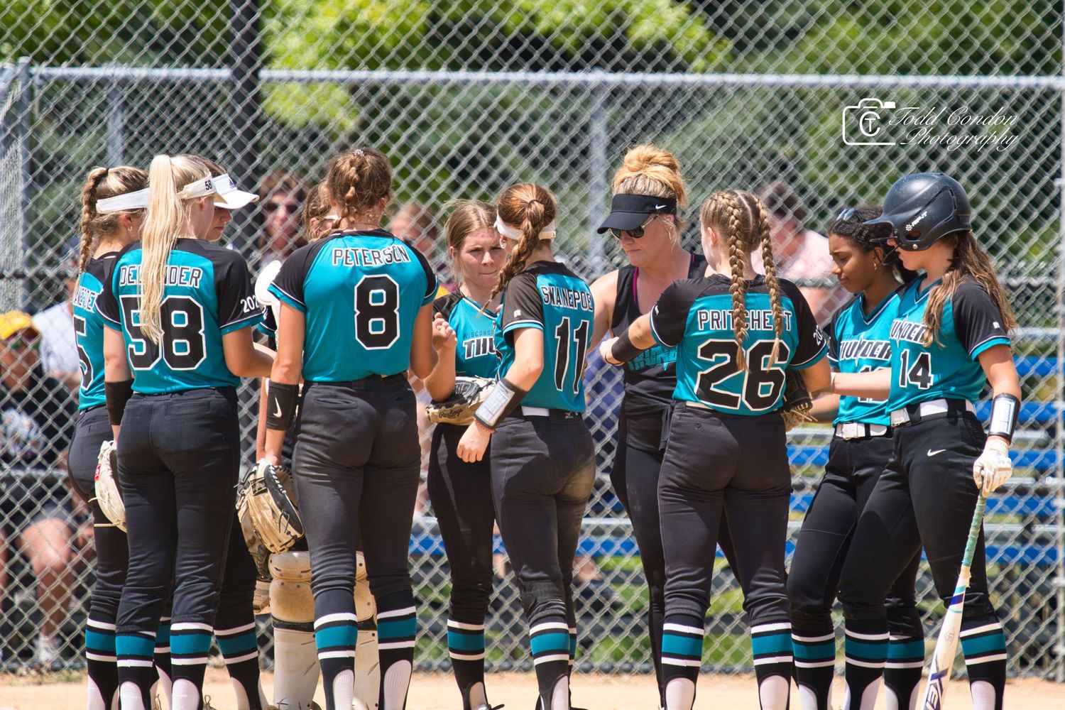 Softball team in teal and black uniforms huddles together on sunny field during game.