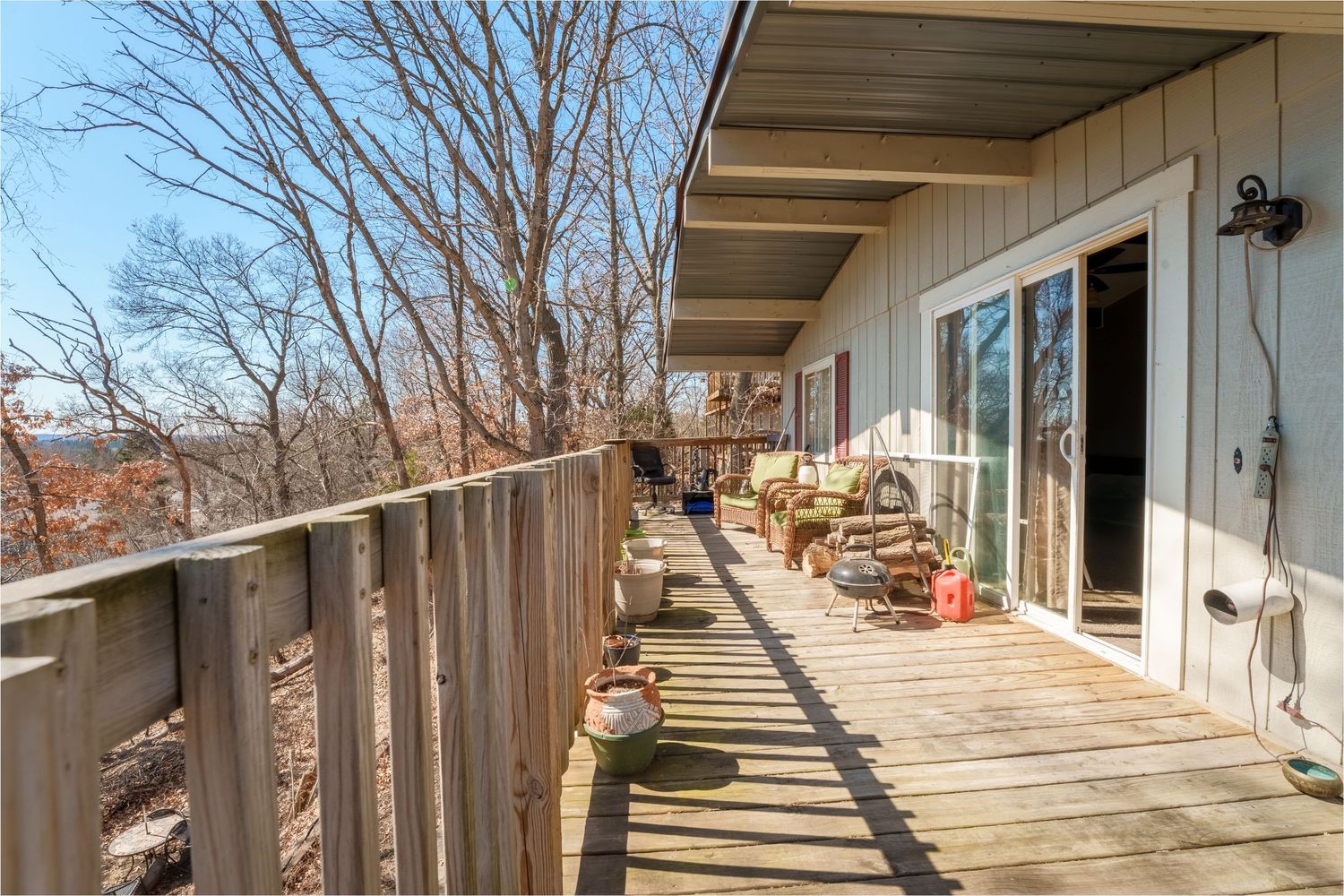 Wooden deck with railing extends along the side of a house on a hillside with bare winter trees in the background.