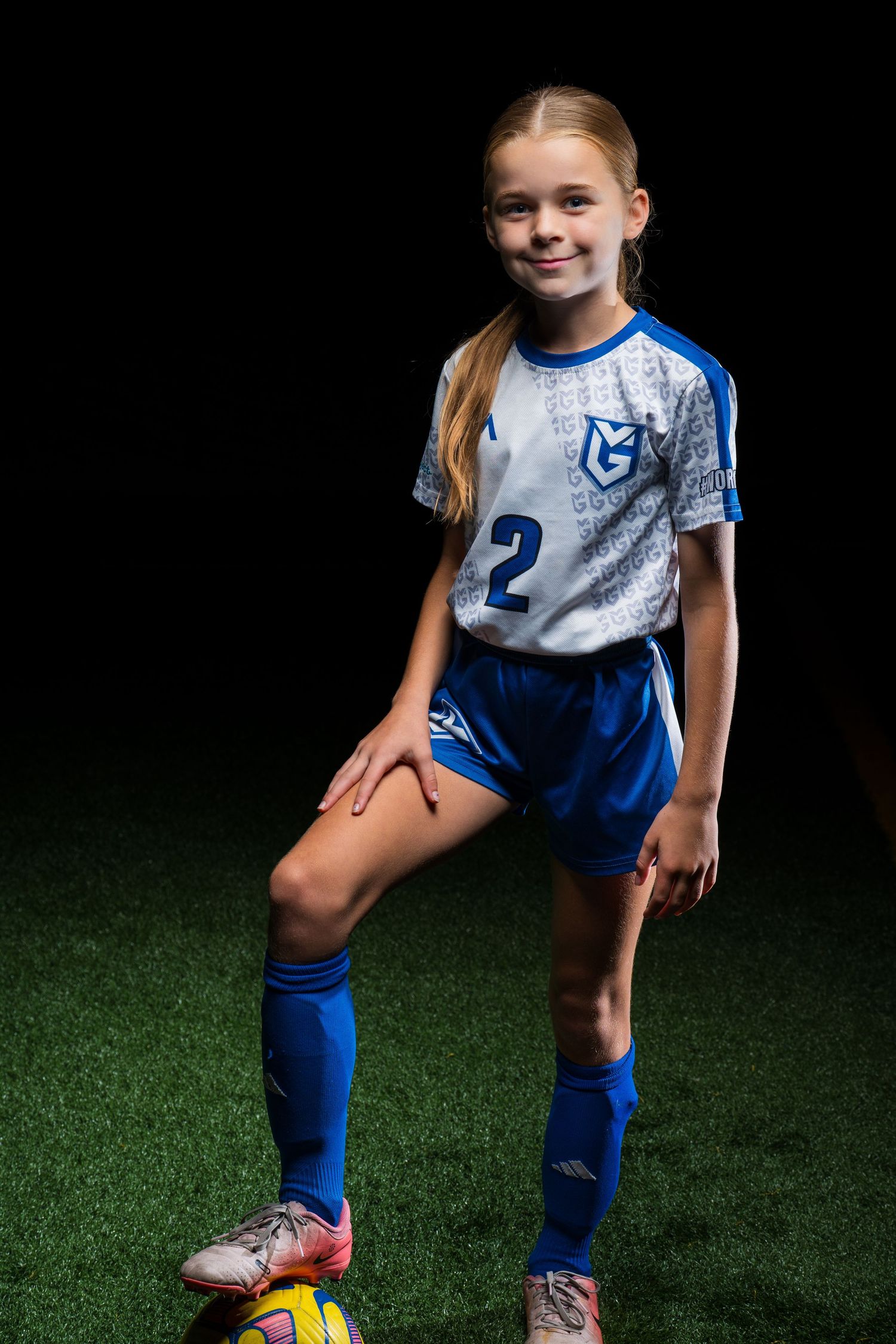 Young soccer player in blue and white uniform poses on dark turf field with cleats and knee socks.