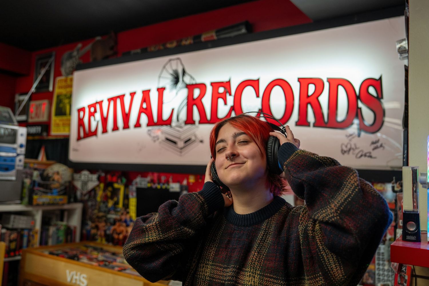 A customer wearing headphones smiles while browsing inside Revival Records store with illuminated signage visible.