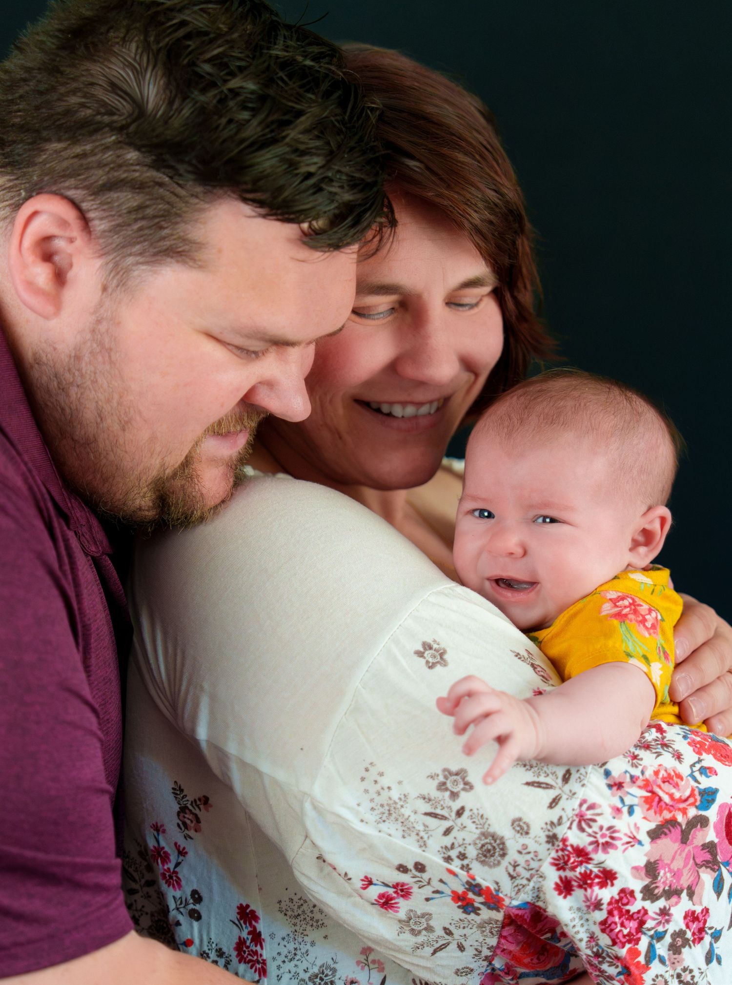 Happy family portrait with parents smiling at their newborn baby dressed in yellow floral outfit.