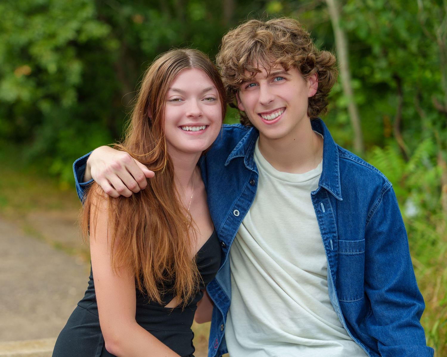 Two smiling teenagers pose together outdoors on a sunny day, girl with long red hair and boy with curly brown hair.