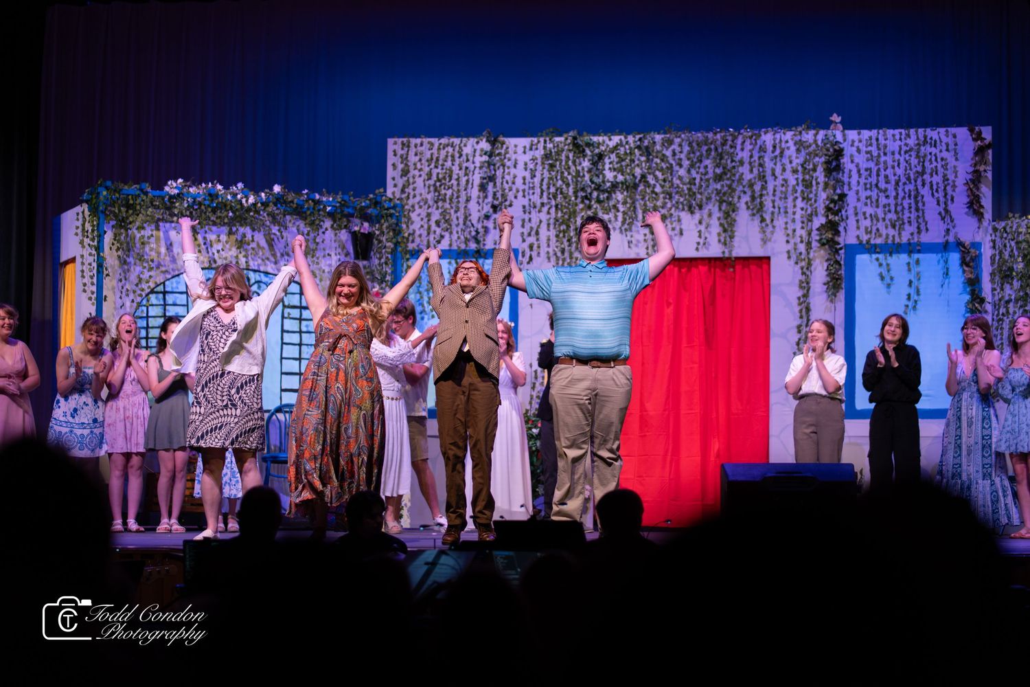 Stage performers in period costumes stand together during a theatrical production with colorful lighting and set pieces.