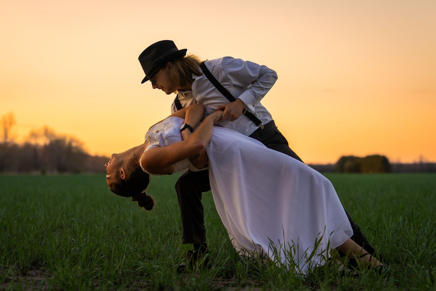 Silhouetted couple embraces in a romantic dance pose against a golden sunset in a grassy field.
