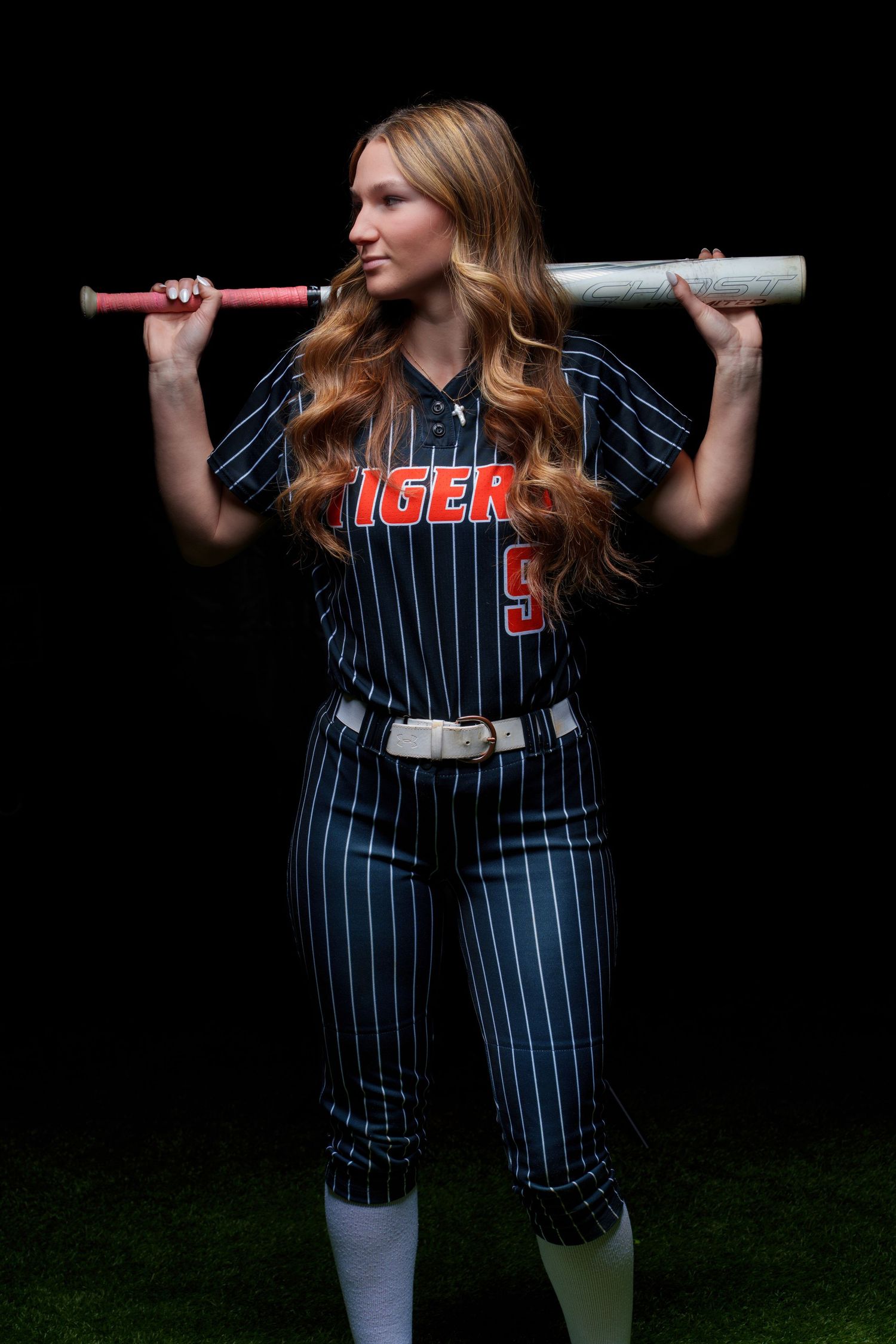 Female softball player in Tigers pinstripe uniform holding bat across shoulders against dark background.