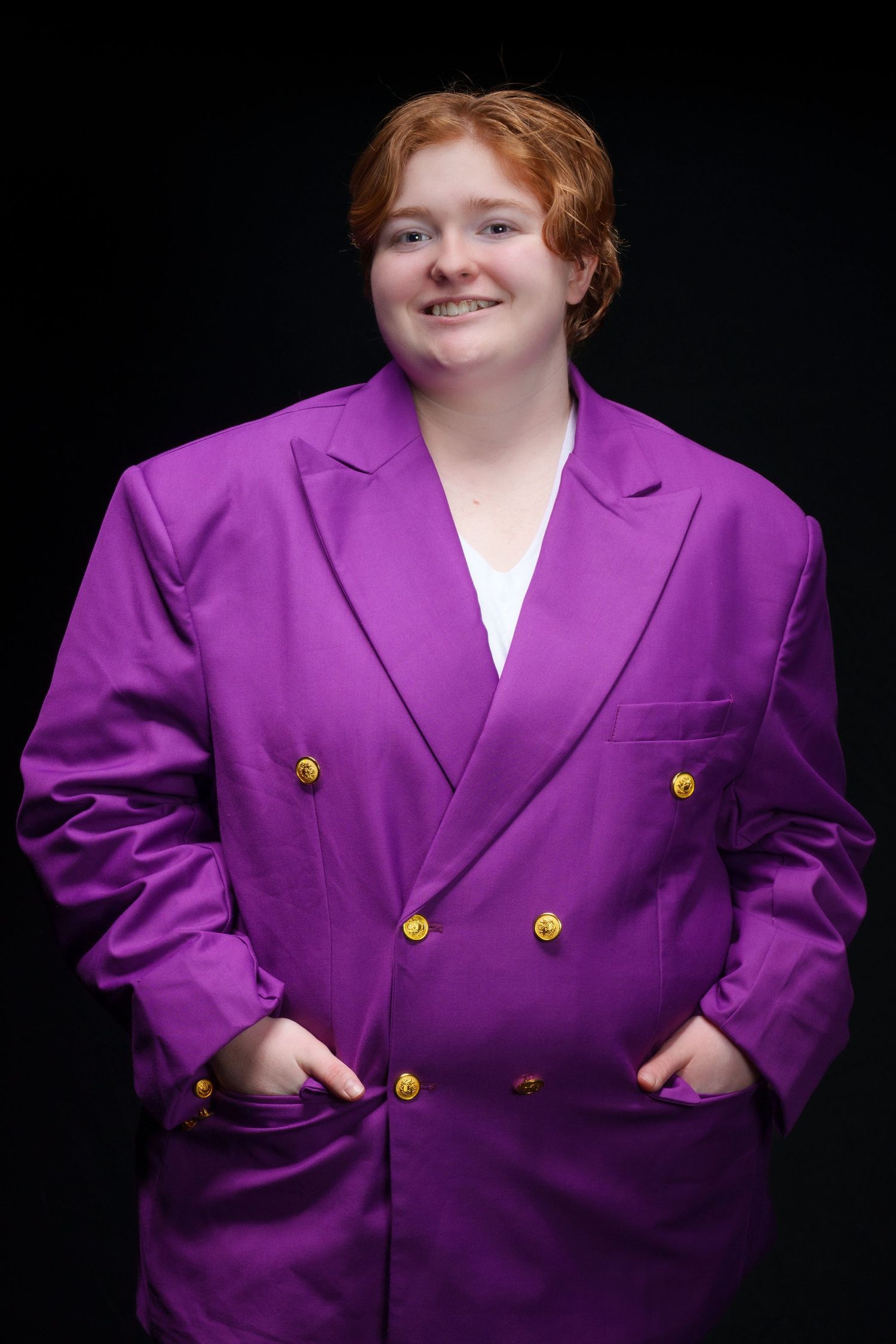 A young person with red hair smiles wearing an oversized bright purple double-breasted blazer with gold buttons on black background.