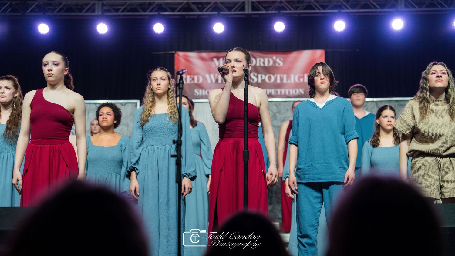 Performers in colorful dresses and outfits stand together on stage under blue lighting during a musical performance.