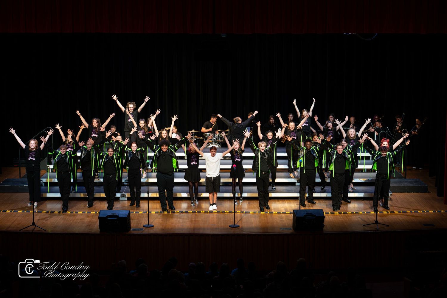 Dancers perform a synchronized routine with raised arms on a dimly lit stage during a recital performance.