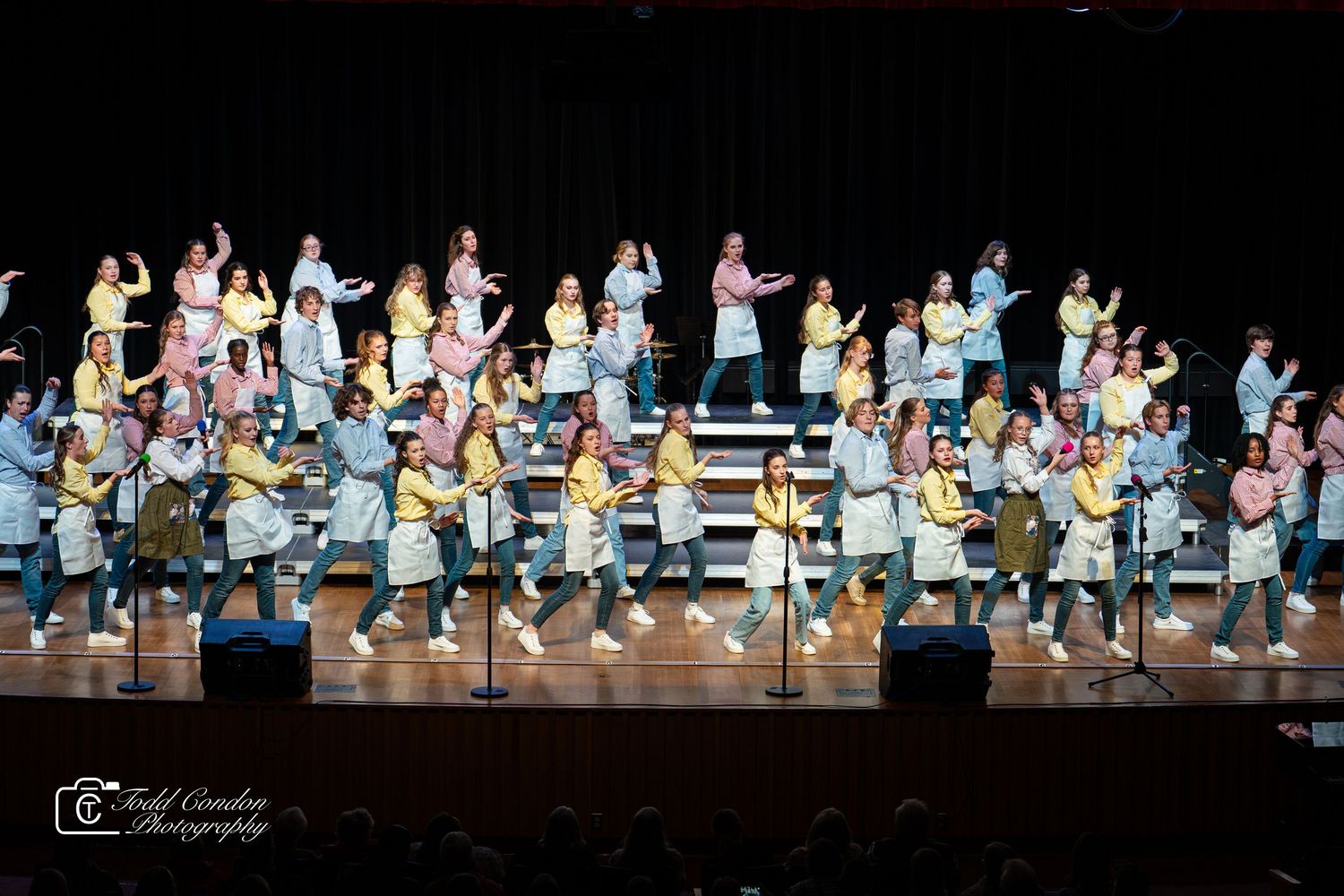 Large group performs synchronized dance routine on risers during stage performance in light blue and white costumes.