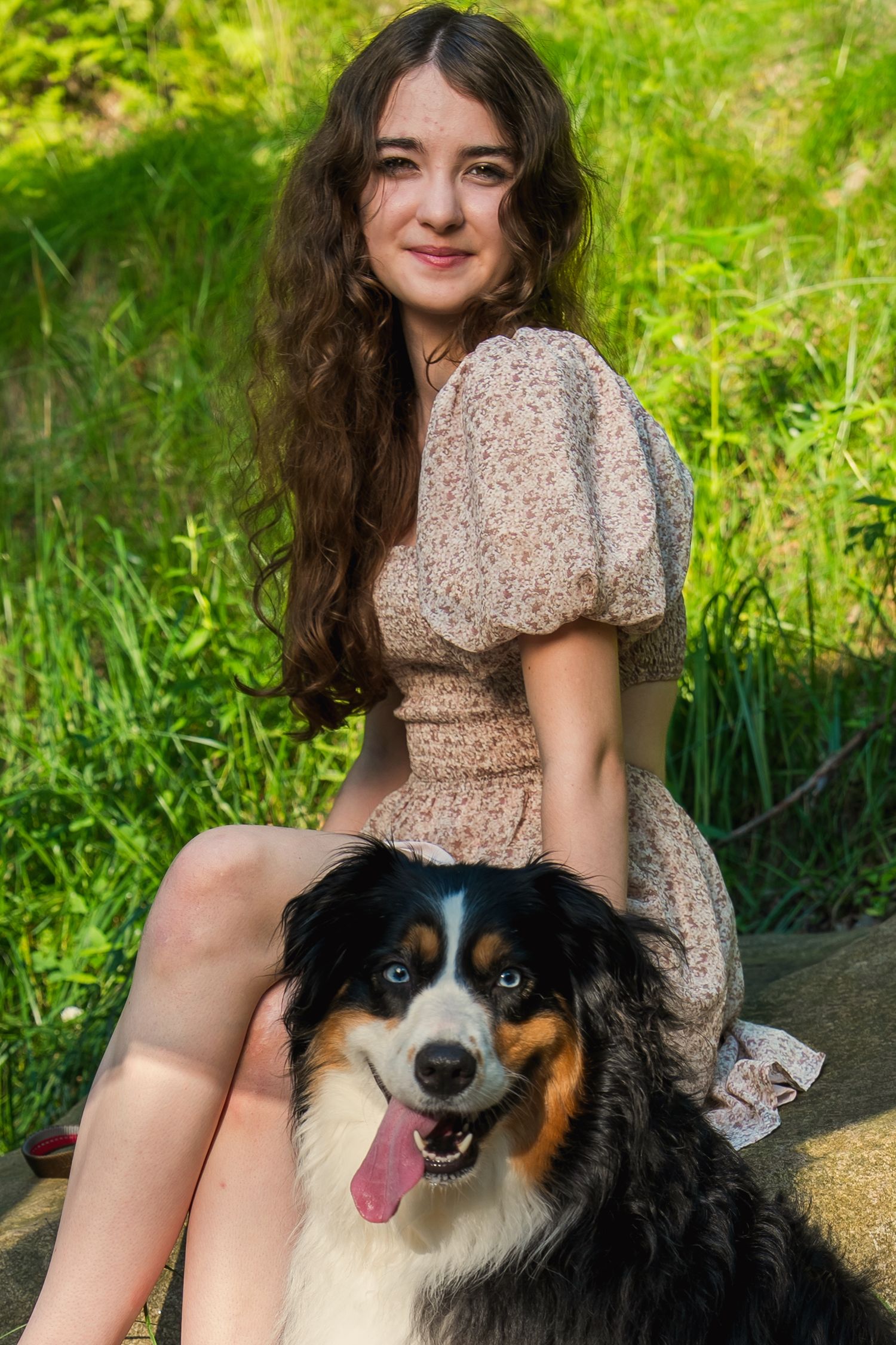 A happy Australian Shepherd dog sits with someone wearing a beige floral dress in a sunny garden setting.