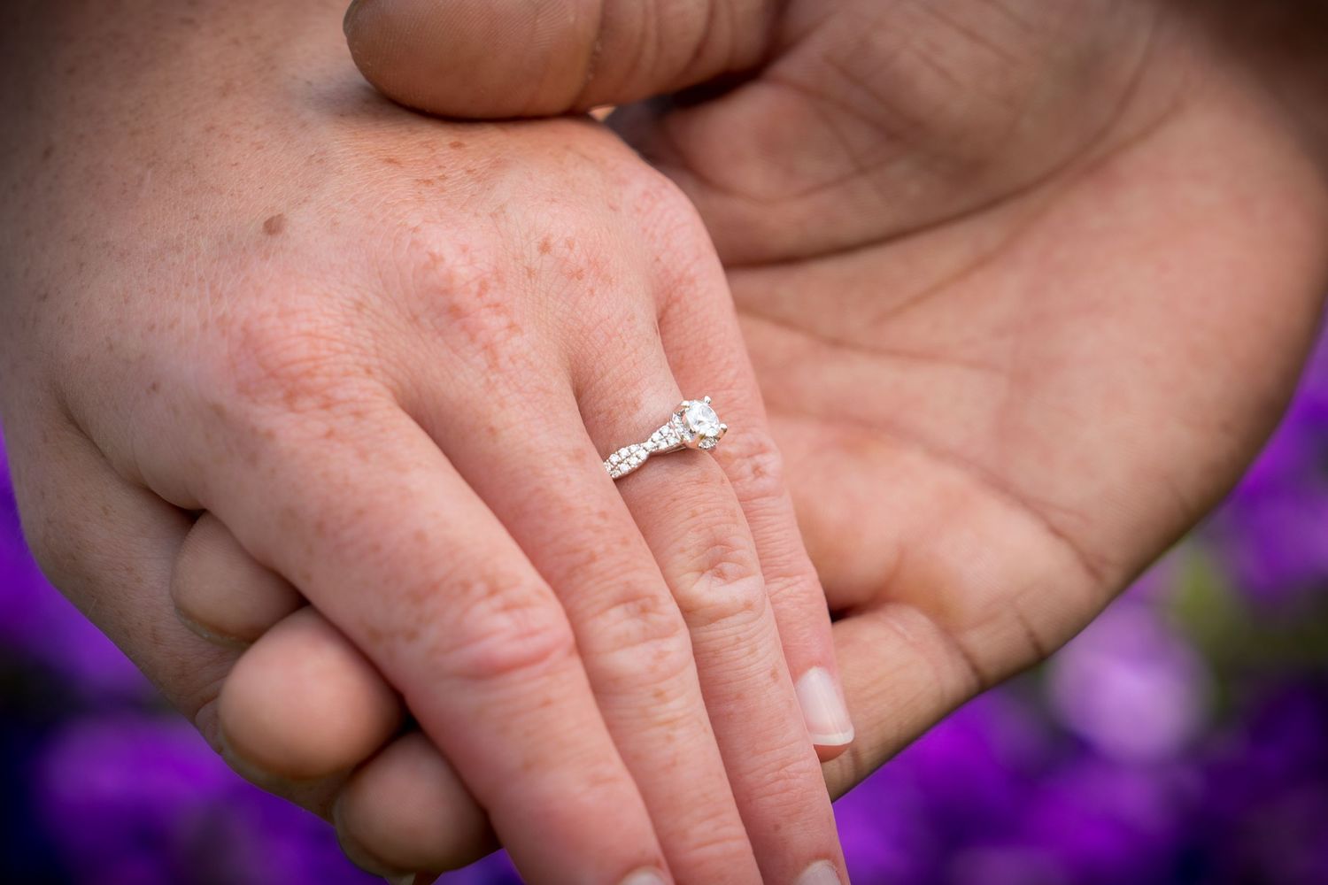 A diamond engagement ring sparkles on a hand with purple flowers in the background.