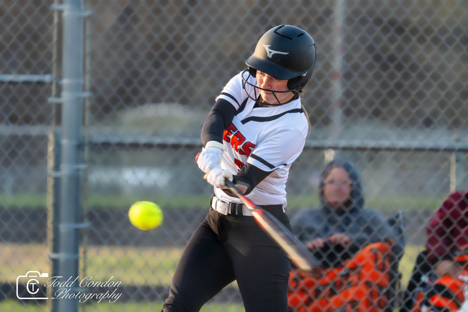 Softball player in white uniform swings bat at incoming yellow ball during competitive game.