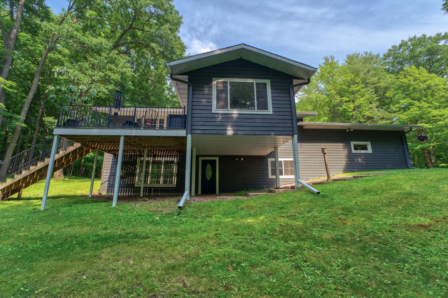 Gray two-story home with large upper deck, green lawn, and surrounding trees on a sunny day.