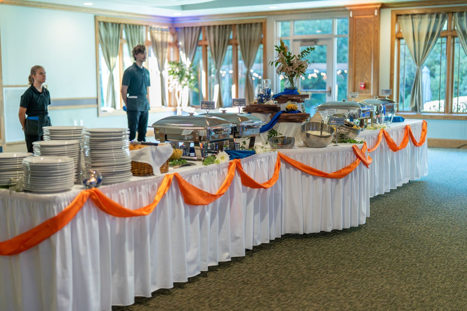 Long banquet tables with white tablecloths and orange draping decorations line a carpeted event venue space.