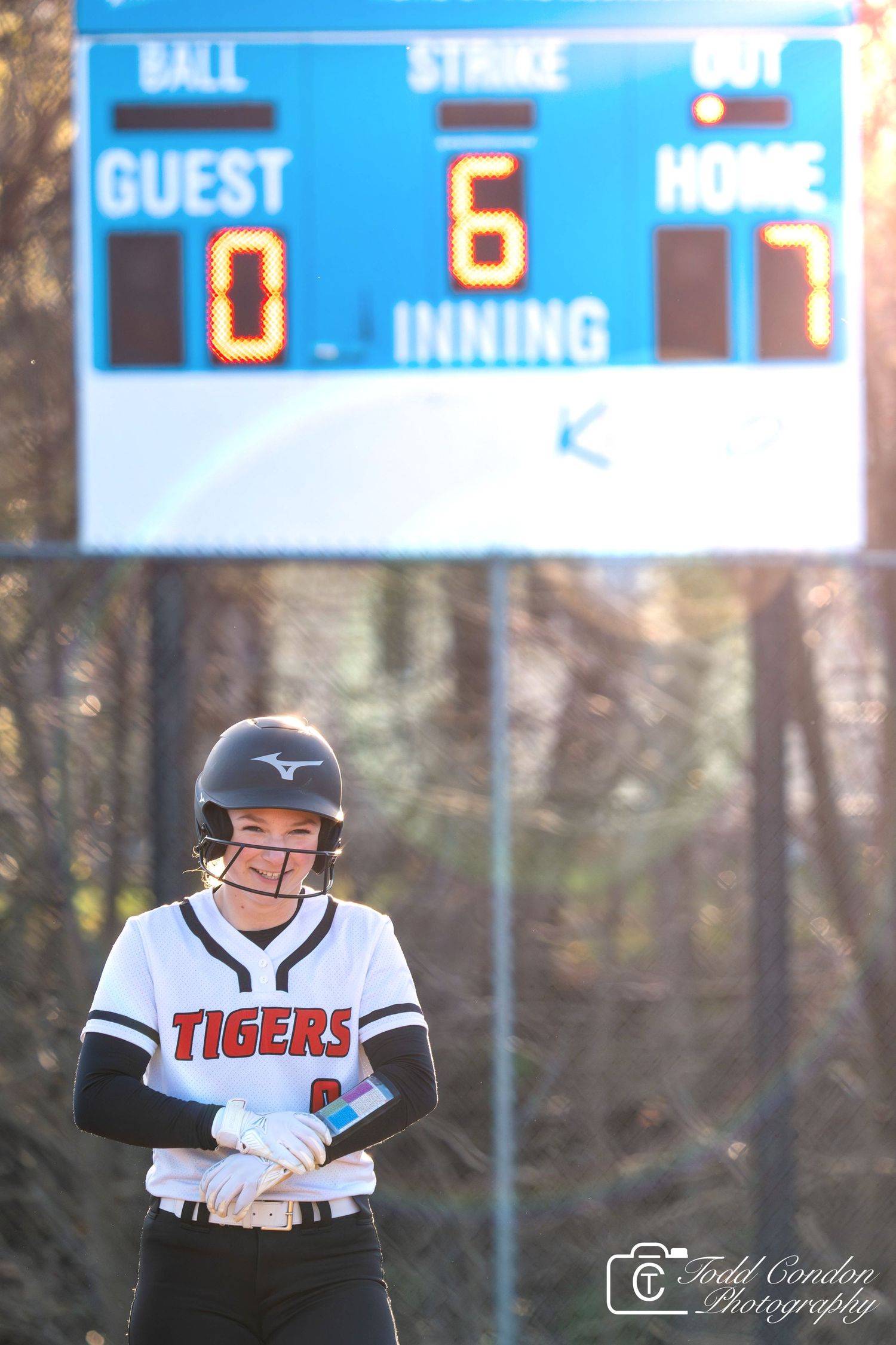 Baseball player in Tigers uniform stands ready at bat during evening game with scoreboard visible in background.
