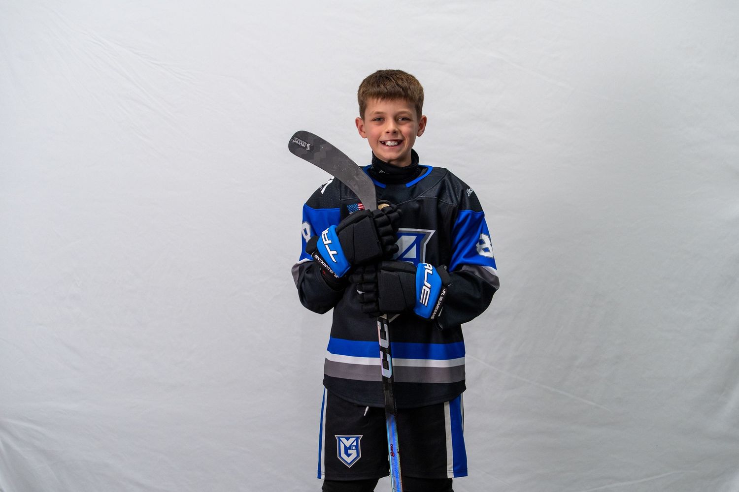 Young hockey player wearing blue and black jersey and protective gear poses against white background.