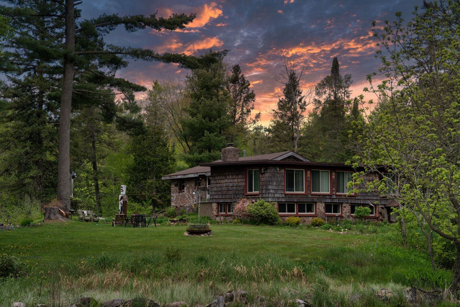 A rustic log cabin nestled among pine trees at sunset with dramatic orange and purple clouds in the sky.