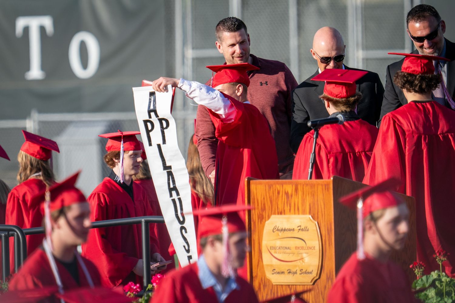 Graduation ceremony with students in red caps and gowns, one person holding an applause sign at podium.