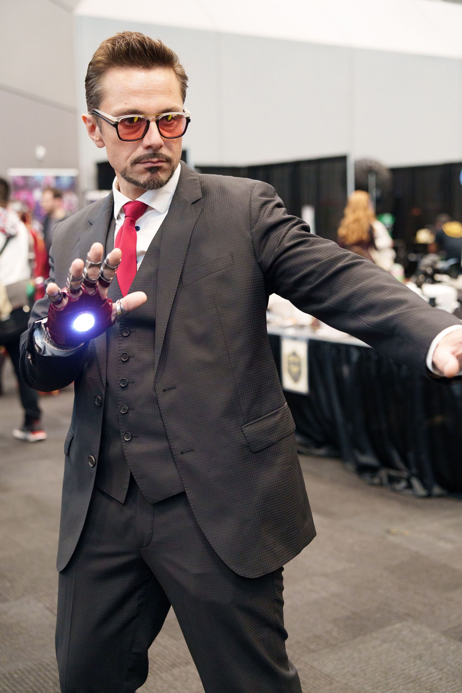 Man cosplaying as Tony Stark in gray suit, red tie, tinted glasses, and glowing Iron Man gauntlet at comic convention.