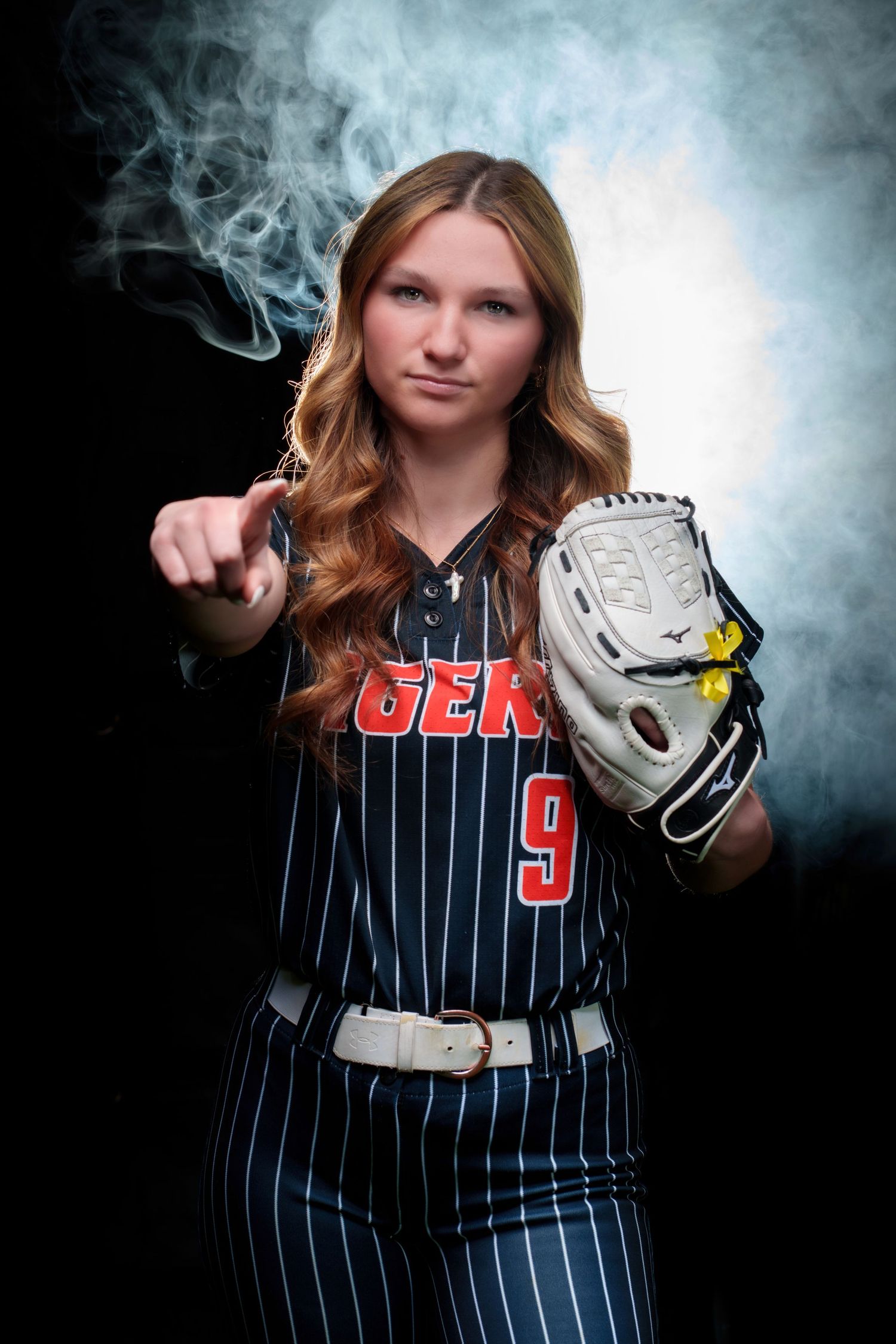Female softball player in pinstripe uniform #9 pointing at camera while holding a fielding glove, dramatic smoke background.