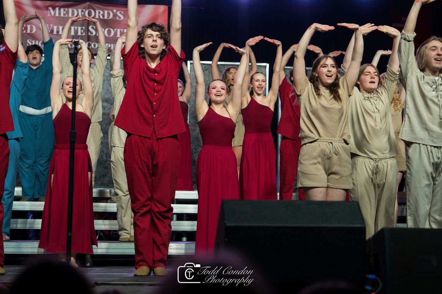 Performers in red and beige costumes stand on risers with raised arms during a stage performance.