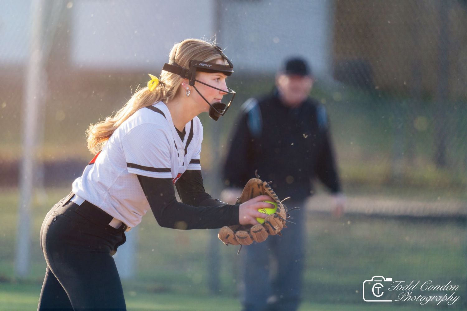 A softball player in black uniform swings the bat during a game at sunset.