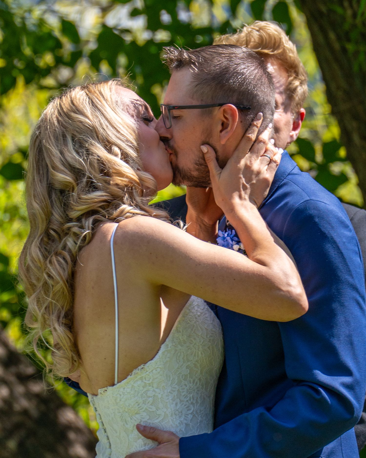 A couple shares a romantic kiss outdoors in a garden setting, with the bride in white and groom in navy blue.