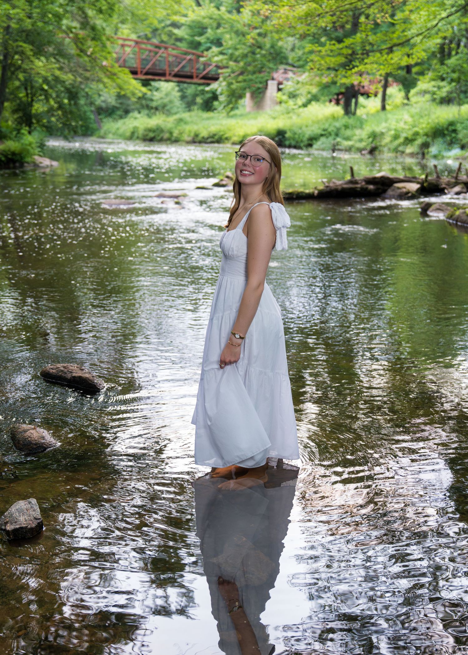 A figure in a white dress stands gracefully in a flowing stream surrounded by lush greenery and rocks.