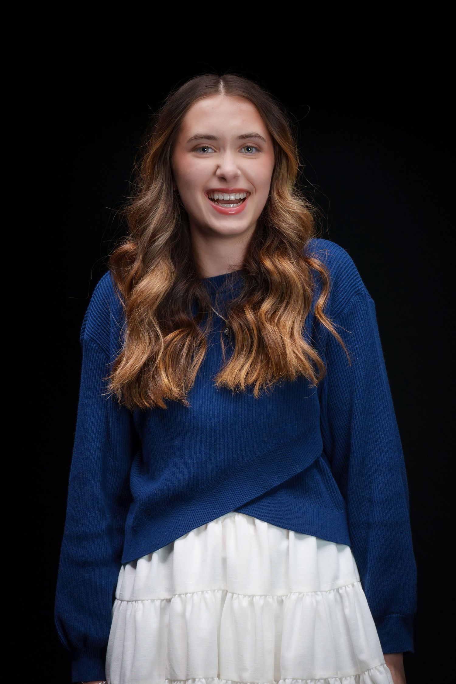 Smiling young woman with wavy auburn hair wearing a blue sweater and white skirt against a dark background.