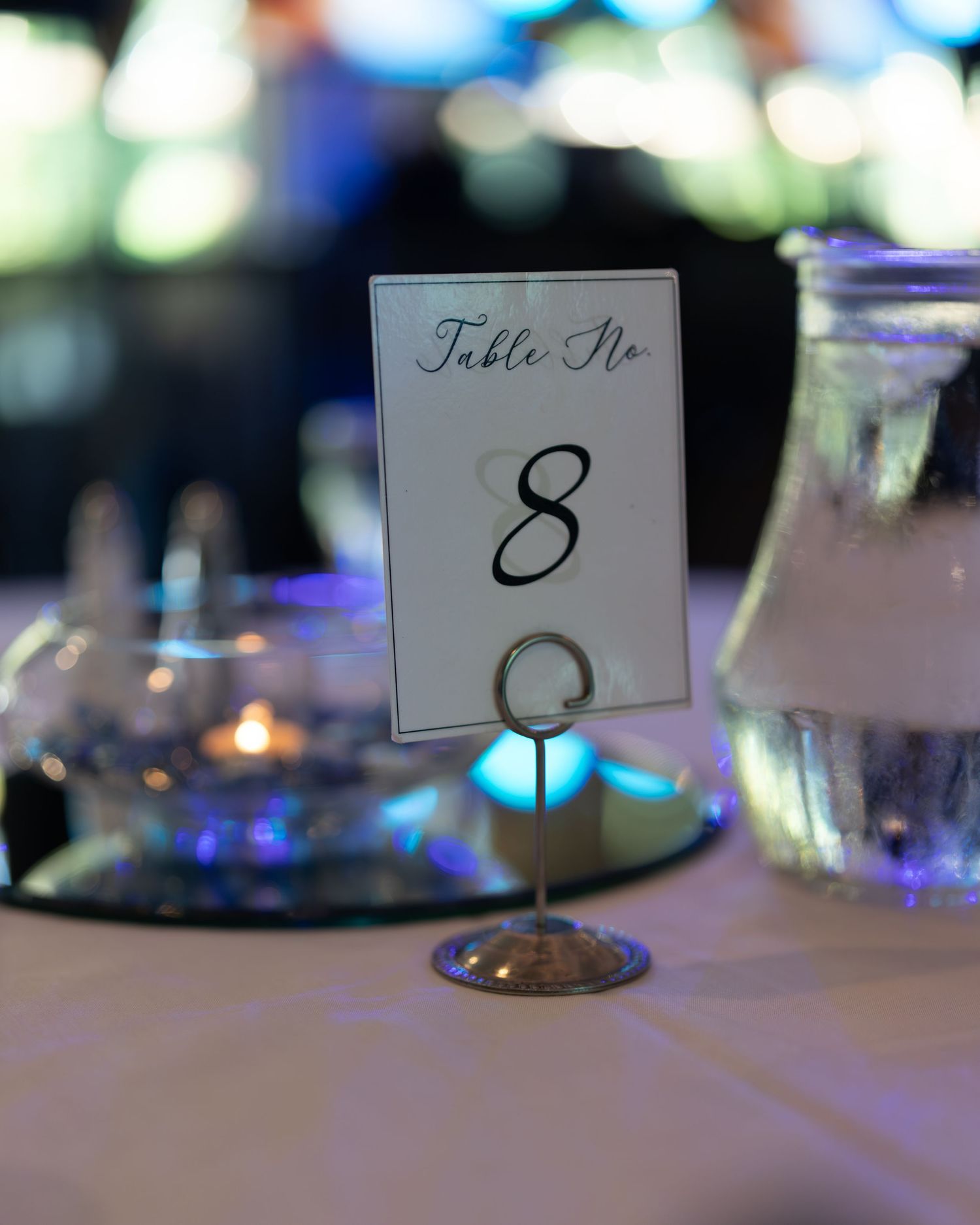 Elegant table number card with calligraphy number 8 displayed at a formal event with bokeh lighting in background.