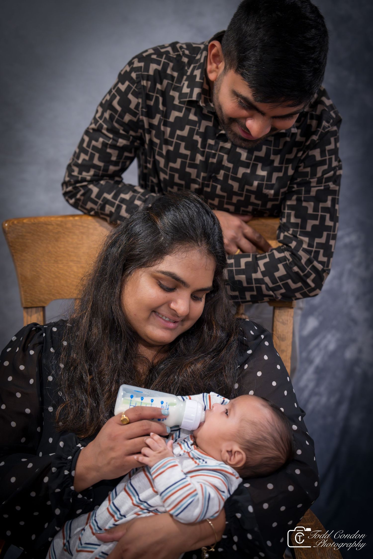 A tender family moment as parents bottle feed their newborn baby in a studio portrait setting.