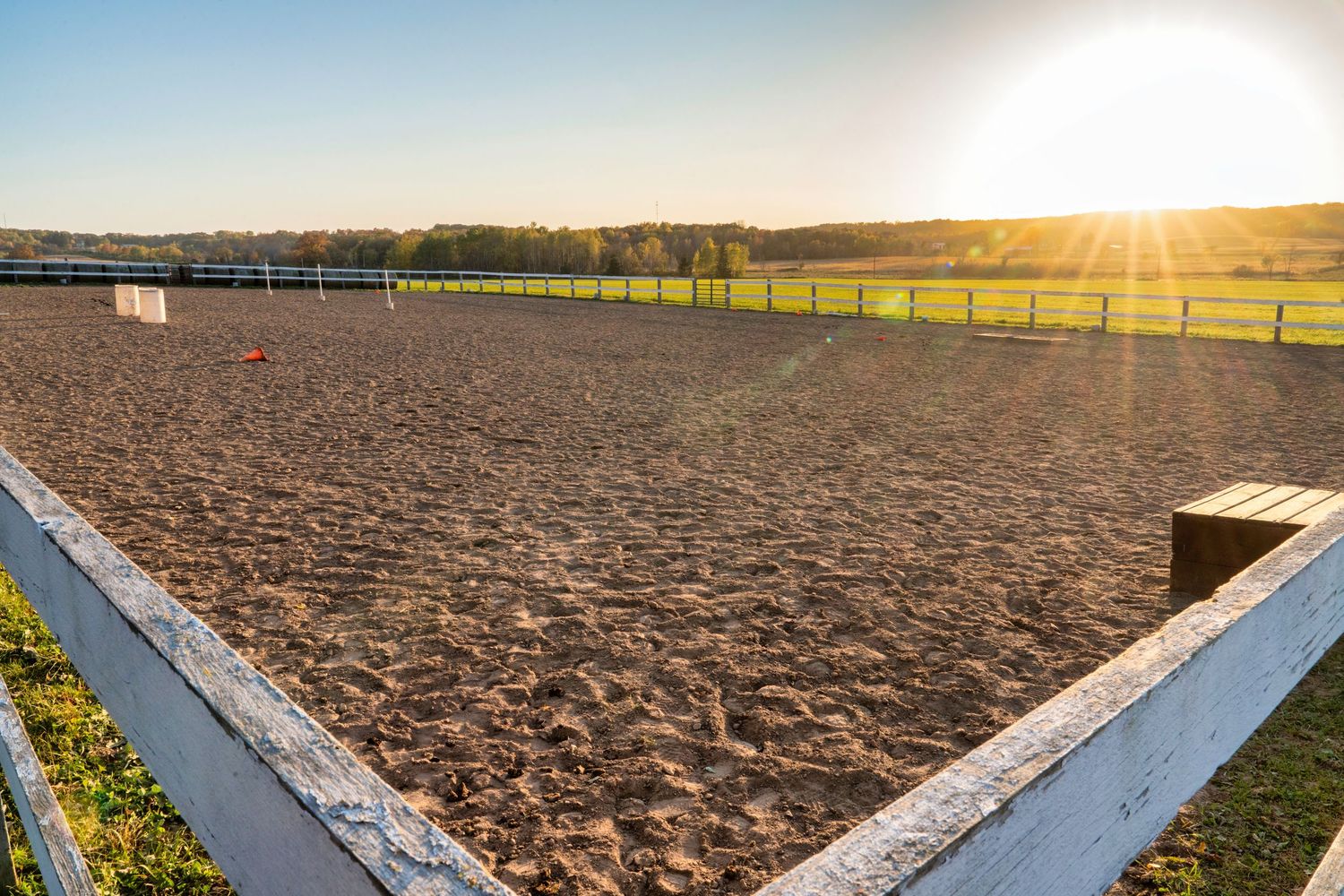 Empty riding arena with white fencing and freshly groomed sand surface at sunset.