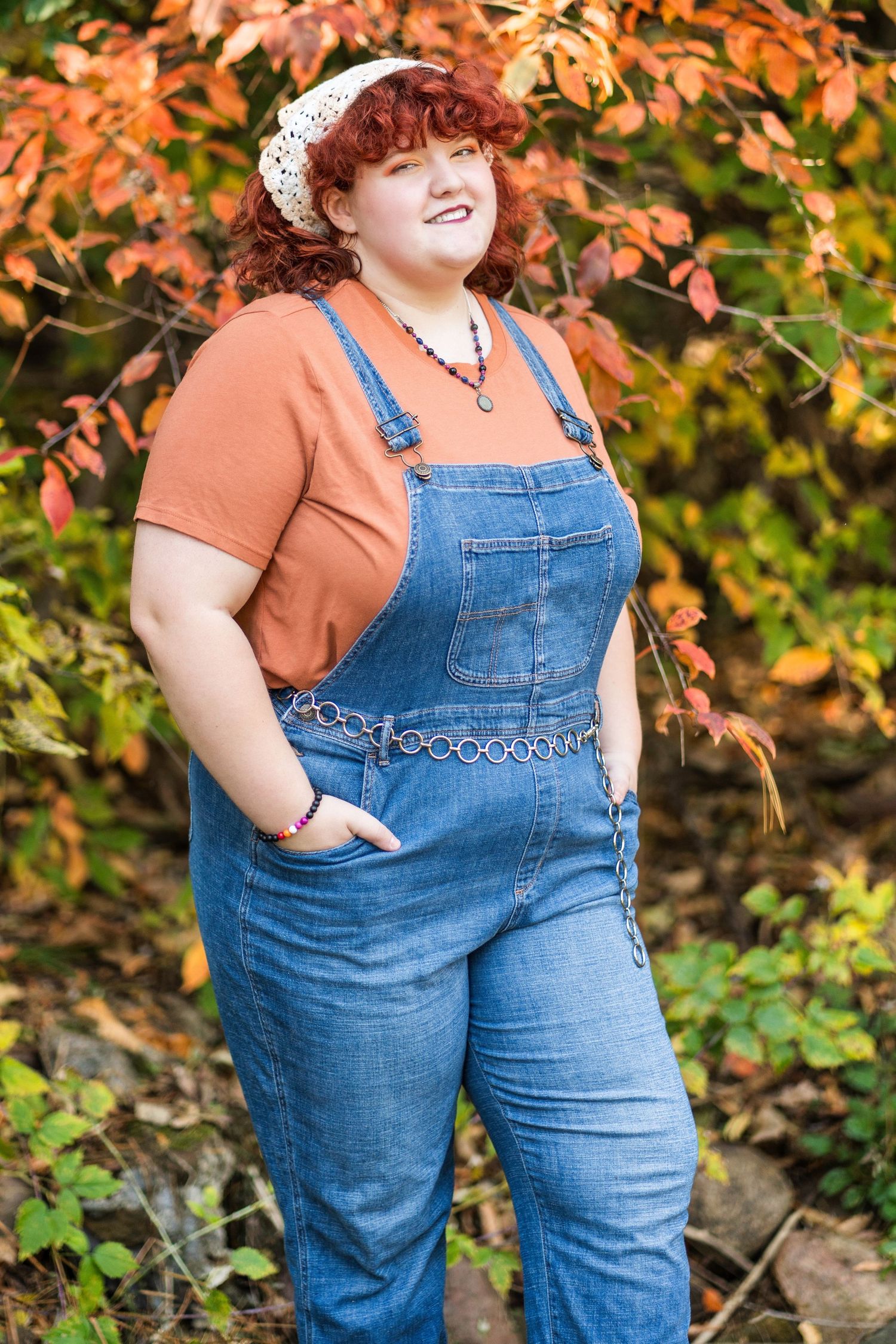 Person wearing blue denim overalls poses outdoors against autumn foliage in orange and green tones.