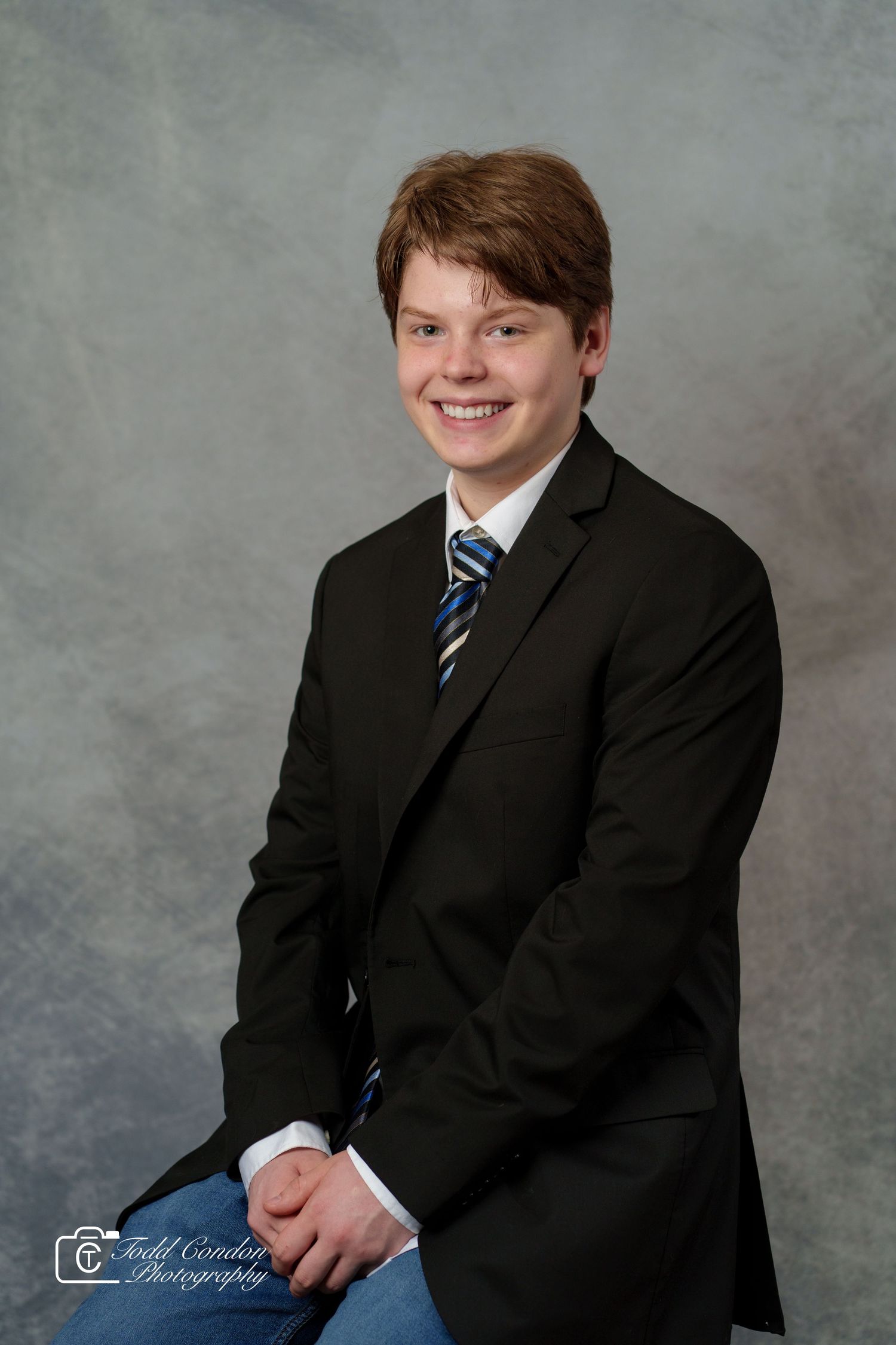 Smiling teenage boy in black blazer, striped tie, and jeans poses for a professional portrait photo.