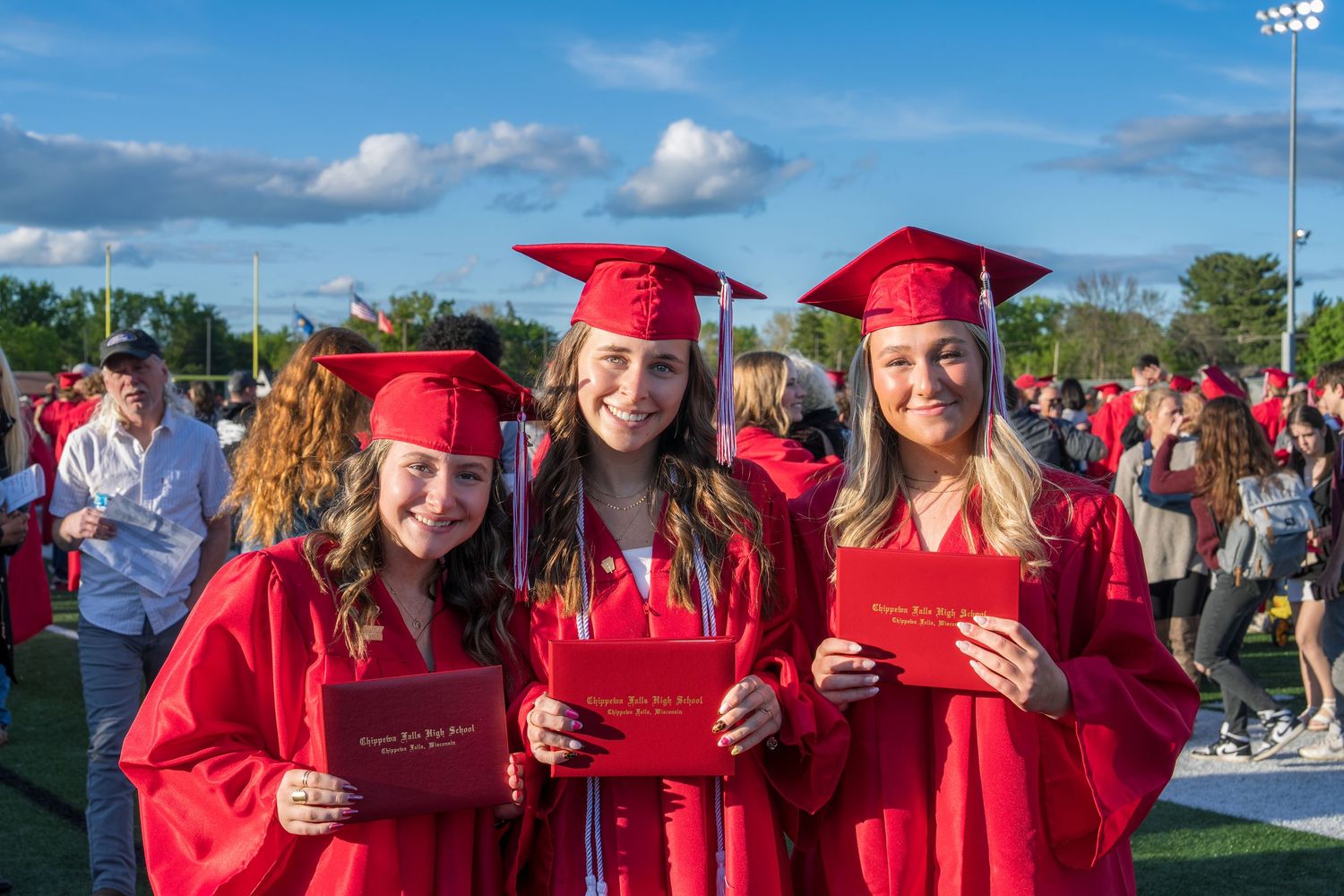 Three smiling female graduates in red caps and gowns holding diplomas outdoors at a graduation ceremony.