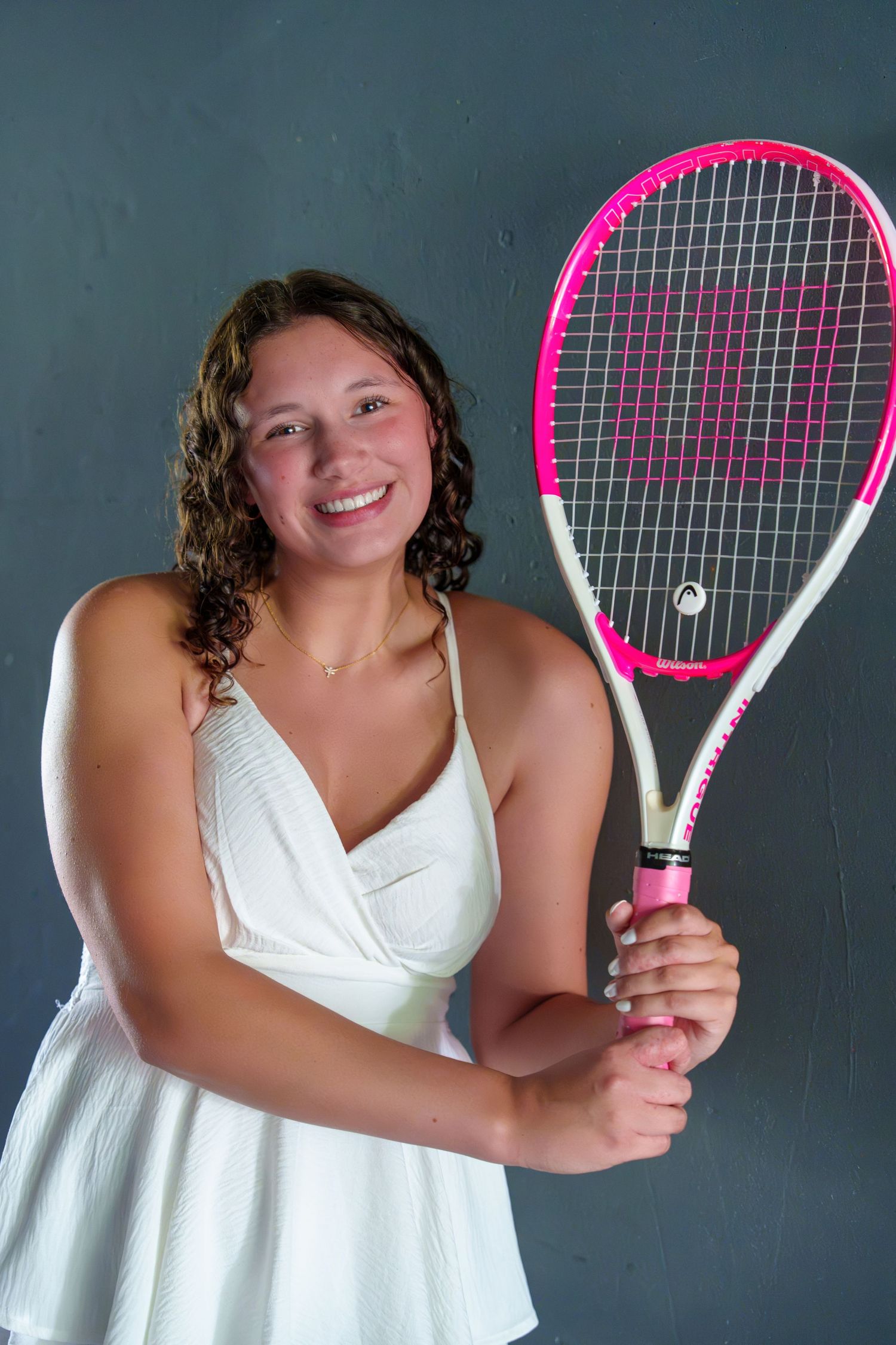 Smiling young woman in white dress holding a pink and white Wilson tennis racket against a gray background.