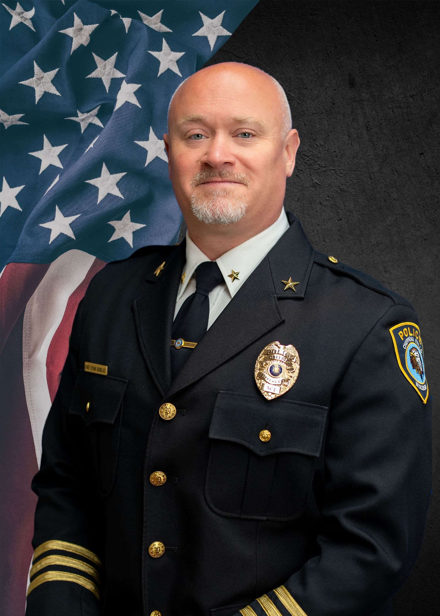 A bald police officer in dress uniform with badge and stars poses formally in front of an American flag backdrop.