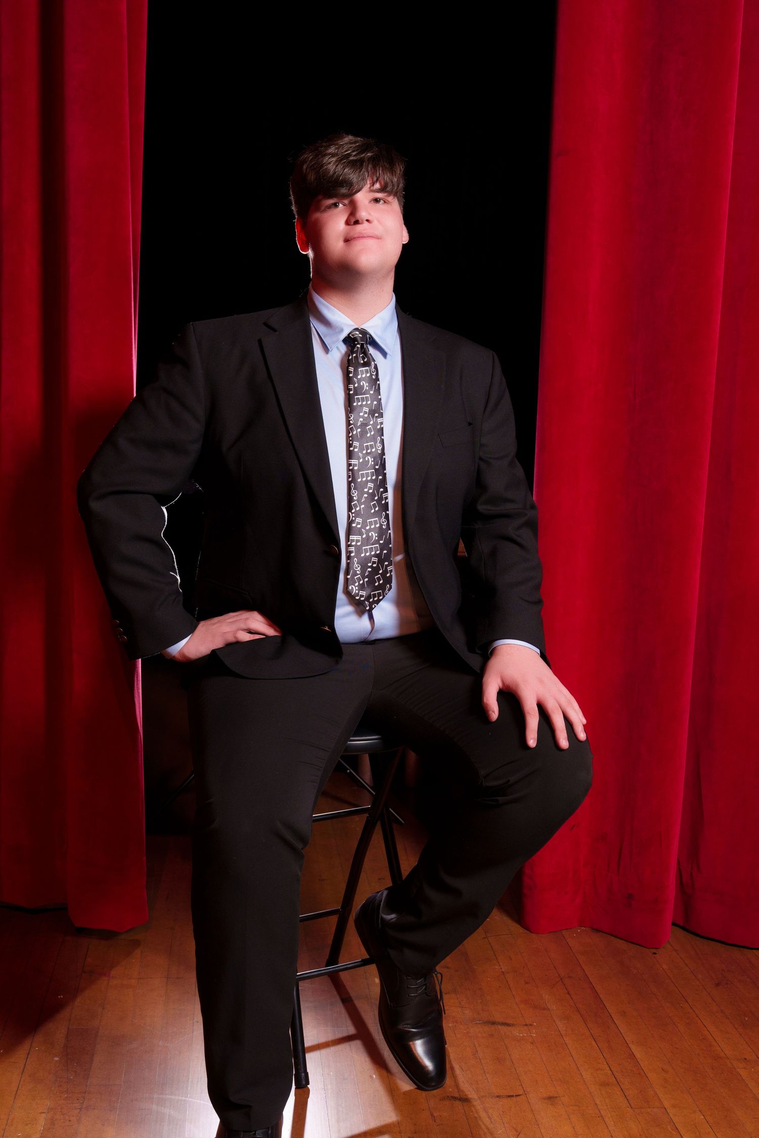 Young man in black suit and patterned tie seated on stool before red stage curtains on wooden floor.