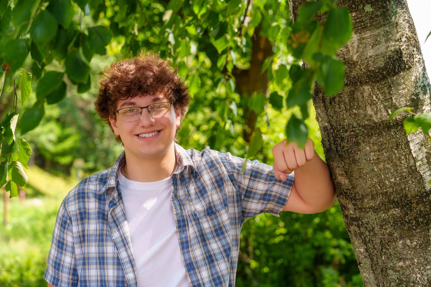 Smiling teenage boy with curly hair and glasses leaning against a birch tree in a lush green park.