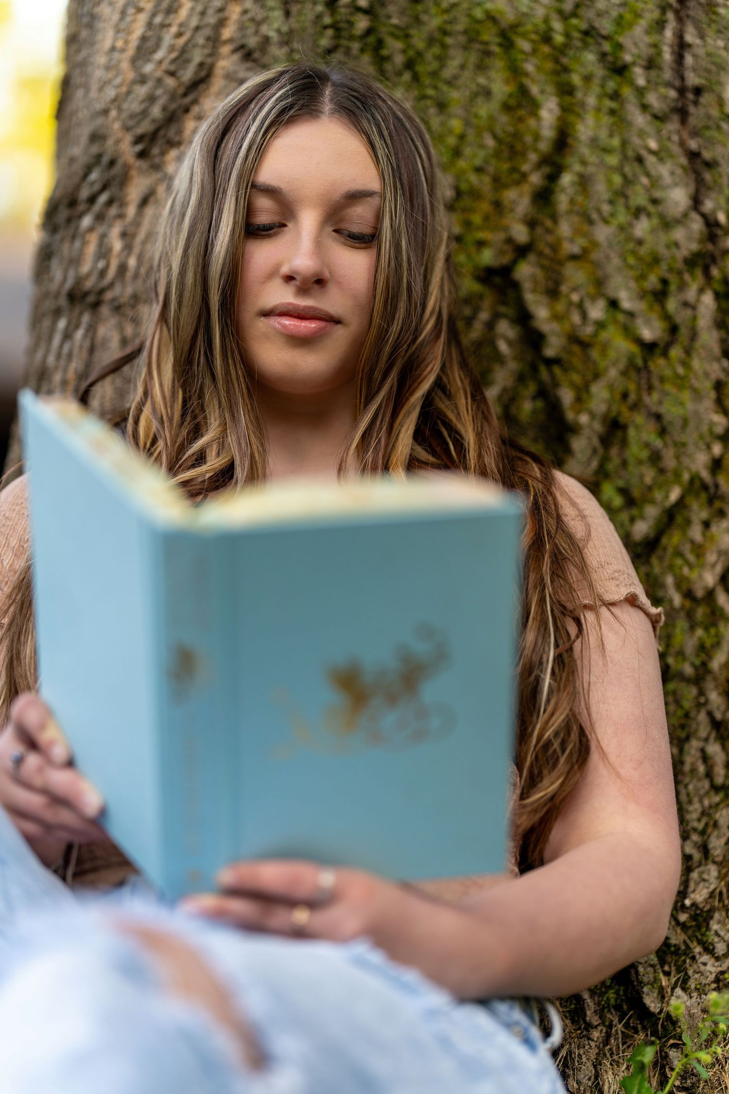 A person with long brown hair reads a light blue book while sitting against a tree trunk in natural lighting.