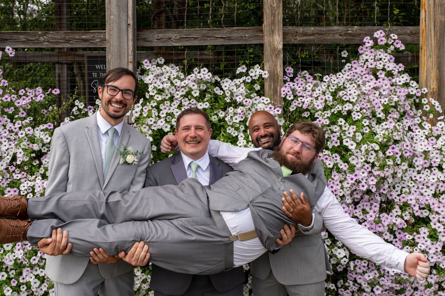 Groomsmen in gray suits pose playfully in a garden setting with white flowers in the background.