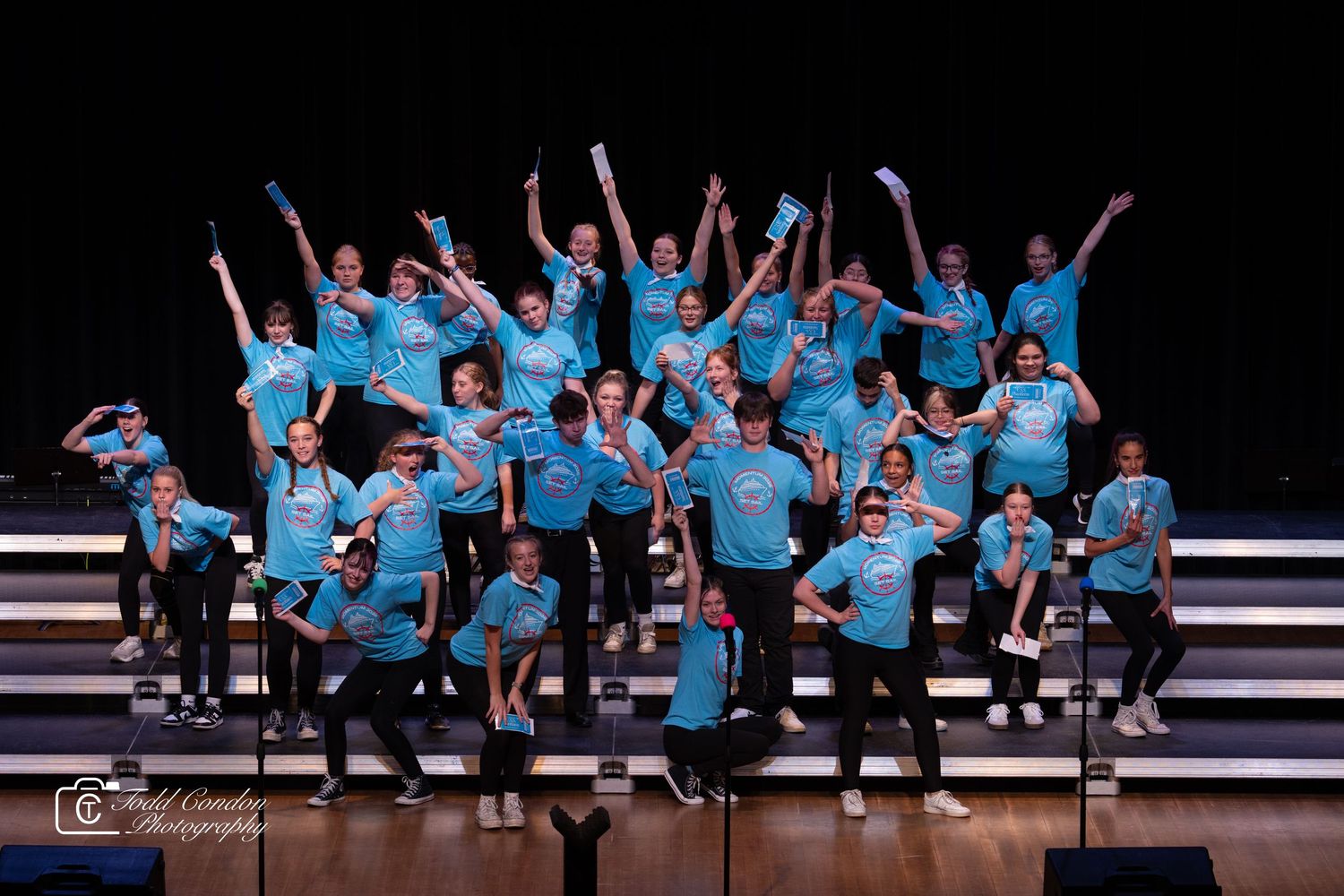 Show choir performers in matching light blue shirts pose energetically on risers during a stage performance.