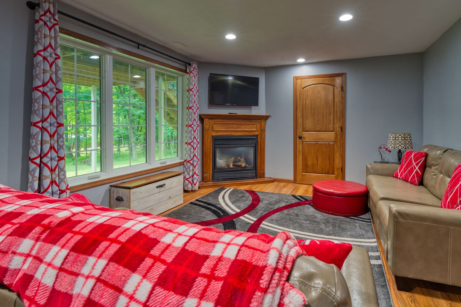 Cozy bedroom with red plaid bedding, corner fireplace, hardwood floors, and large windows overlooking lush greenery.