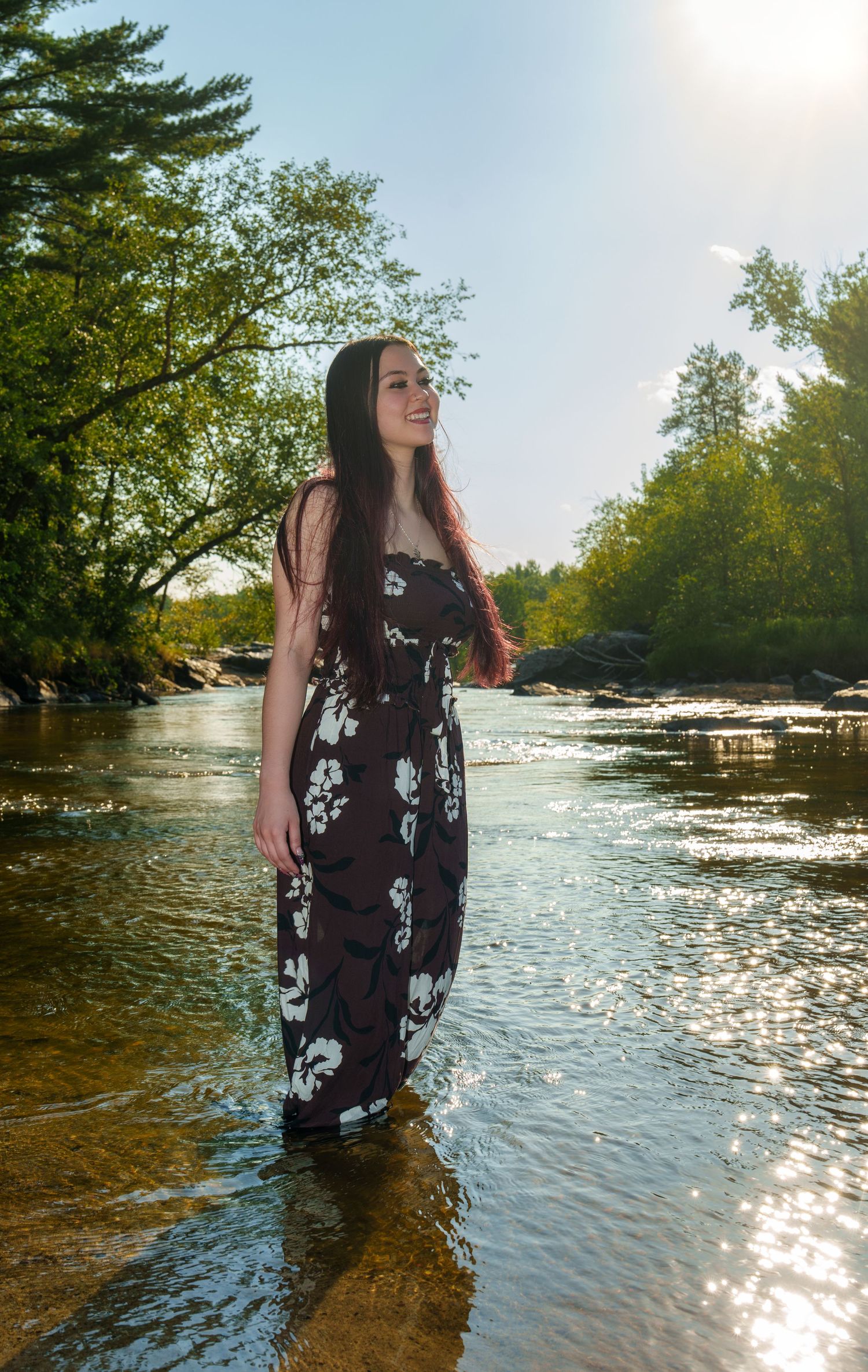 Young woman in floral maxi dress standing in a shallow river, smiling, surrounded by lush green trees.