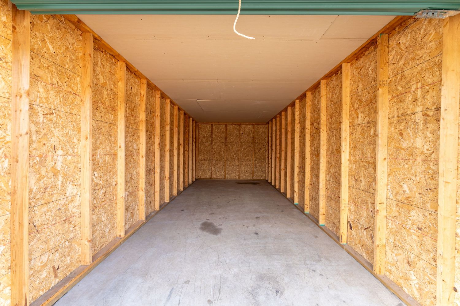 A long wooden hallway with exposed plywood walls and ceiling leads to a bright doorway at the end.