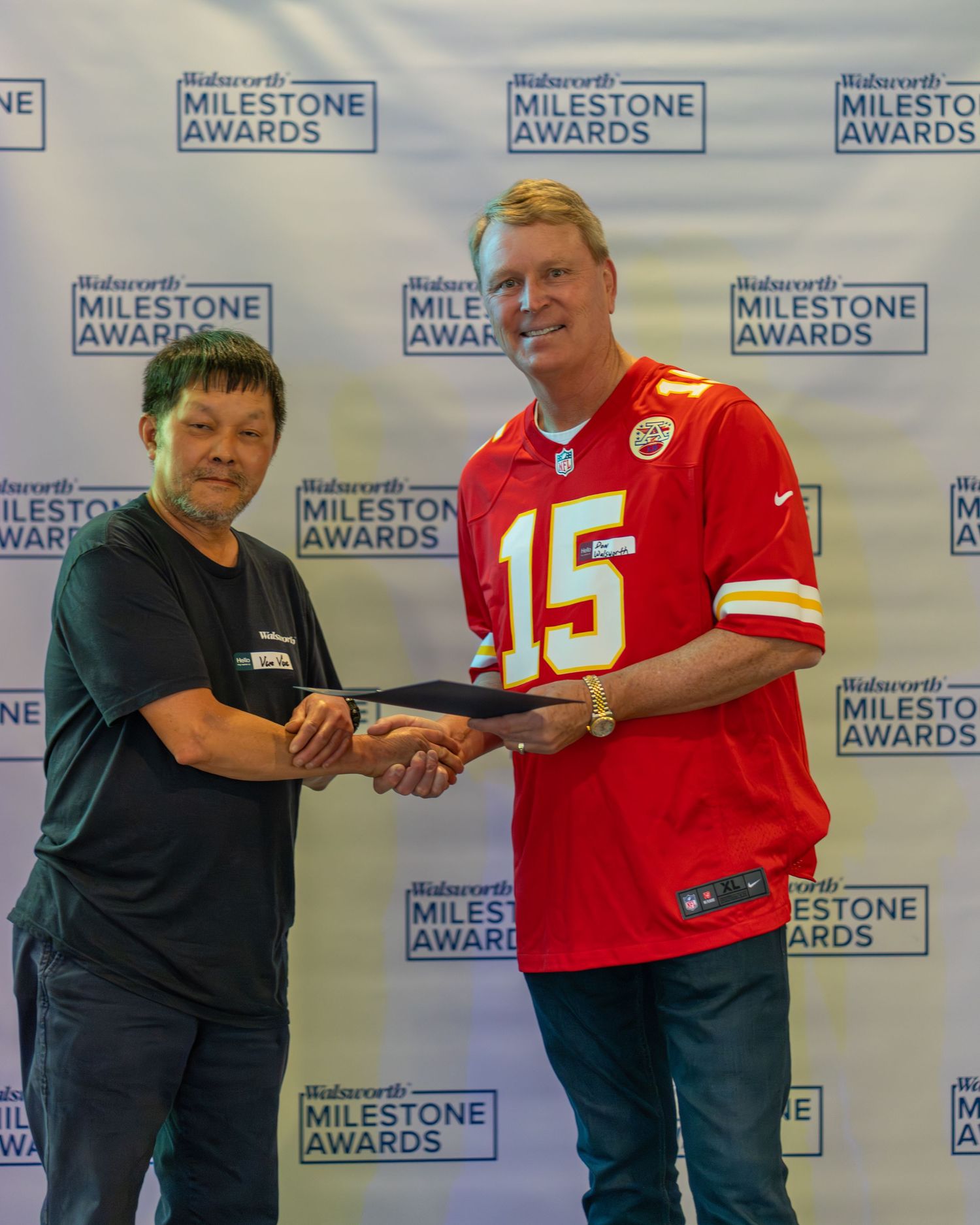 Two people shake hands at an event with Milestone Awards backdrop, one wearing a Kansas City Chiefs #15 jersey.