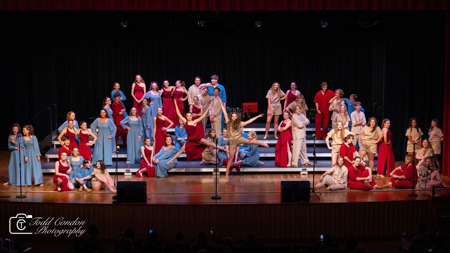 Theater performers in red costumes arranged across a dark stage during a live performance.