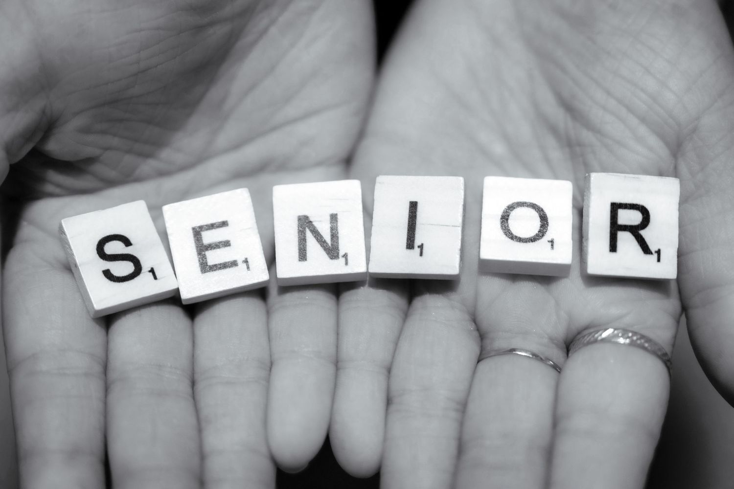 Multiple hands joined together displaying letter tiles spelling out 'SENIOR' in black and white.