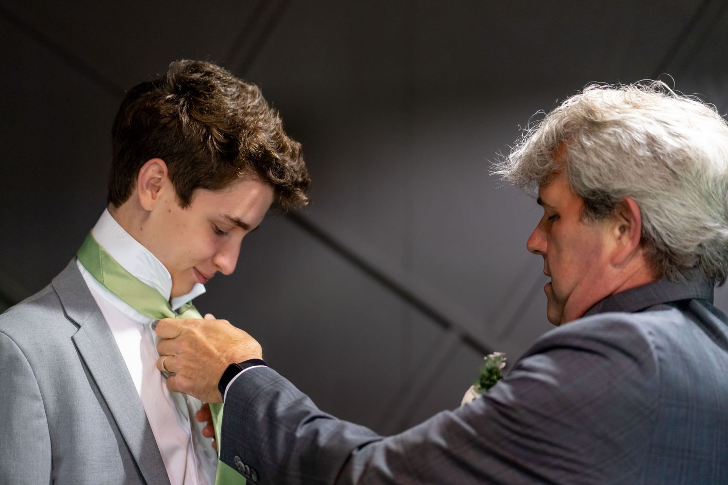 A man in a gray suit gets help adjusting his tie before a formal event.
