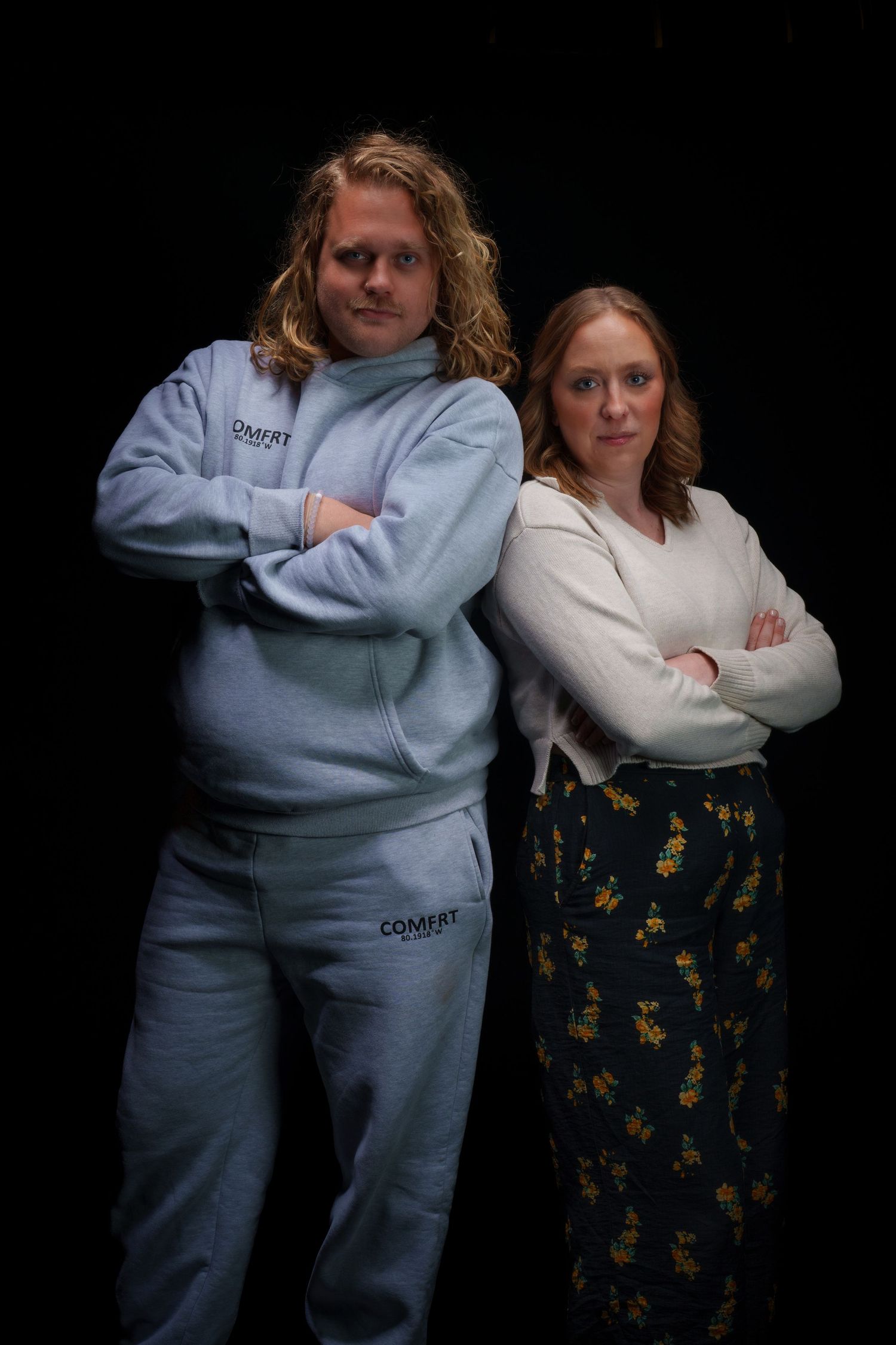 Two people standing back-to-back with arms crossed against a dark background, wearing casual clothing.